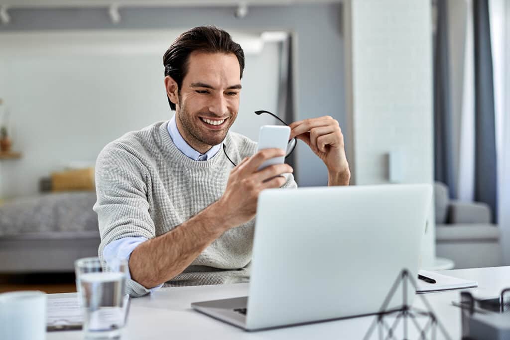 PayFac - Man sitting and smiling at desk with smartphone in one hand and glasses in another hand, sitting infront of laptop