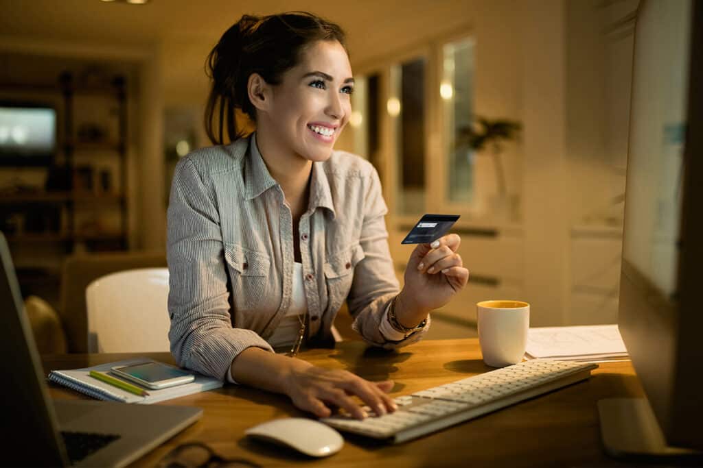 PayFac - woman smiling and sitting down in front of a computer, holding a credit card in one hand, another hand hovering above keyboard